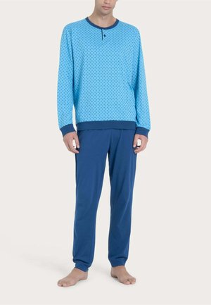 Uomo con maglia a maniche lunghe azzurra chiara con motivo, polsini blu scuro e pantaloni blu scuro tinta unita, in piedi a piedi nudi su uno sfondo uniforme.