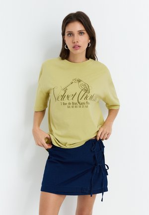 Yellow cotton t-shirt with short sleeves featuring a black printed design. Paired with a navy blue denim skirt with side tie details.