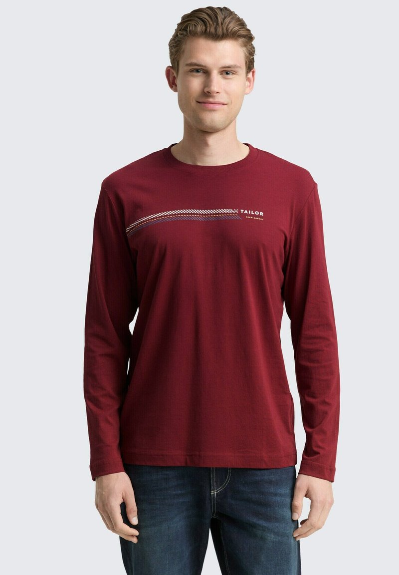 Jonge man in een bordeauxrode longsleeve T-shirt met een diagonale streep en "Tom Tailor" tekst, in combinatie met blauwe jeans, staande en glimlachend.