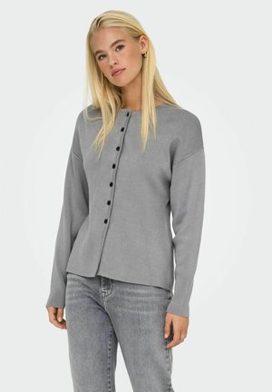 Cardigan long en tricot gris à manches longues avec un col rond, fermeture à boutons sur le devant et coupe décontractée. Porté par-dessus un jean gris clair.