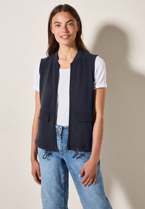 Gilet senza maniche blu navy con chiusura a zip, due tasche frontali e cordoncini regolabili all'orlo; indossato sopra una t-shirt bianca e jeans.