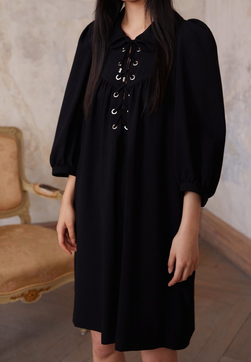 Femme portant une robe noire à manches bouffantes avec un détail de lacets sur le devant, debout à côté d'une chaise vintage tapissée dans une pièce neutre.