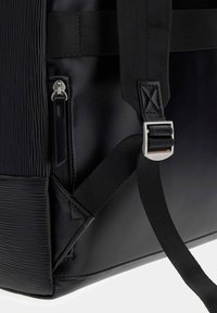 Gros plan d'un coin de sac noir montrant du cuir texturé, une fermeture éclair avec tirette en cuir et une sangle réglable en tissu noir avec boucle métallique.