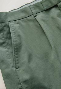 Gros plan de la ceinture d’un pantalon vert montrant les passants de ceinture, la couture latérale et les détails en tissu plissé.