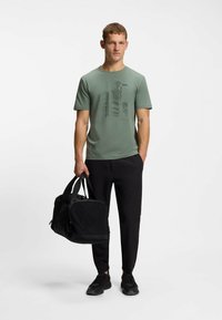 T-shirt verde aderente con design grafico, borsa da viaggio nera, pantaloni neri e sneakers. Tessuto morbido, maniche corte, collo rotondo.