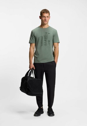 T-shirt verde aderente con design grafico, borsa da viaggio nera, pantaloni neri e sneakers. Tessuto morbido, maniche corte, collo rotondo.