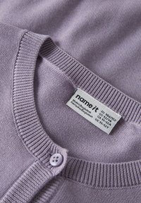 Lys lilla strikket cardigan med knapp og merkelapp som viser størrelse og teksten "Made in Bangladesh".