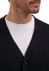 Cardigan di un blu navy scuro con scollo a V, maniche lunghe e chiusura con un bottone, indossato sopra una maglietta bianca a girocollo. Tessuto morbido, dettagli minimi.
