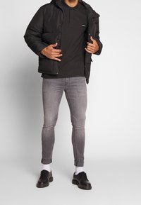 Mustapuffer-impulssi, jossa on huppu, tikattua tekstiä, käytetään mustan polo-paidan päällä. Vaaleanharmaat slim fit -farkut ja mustat kengät täydentävät asua.