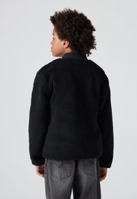 Crna flis jakna s teksturiranom površinom, koja ima visoki ovratnik i duge rukave. Nosena uz sive traperice.