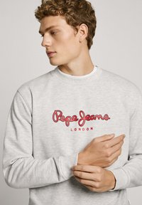 Sudadera gris con un destacado logo rojo de "Pepe Jeans" y el texto "LONDON", cuello redondo y puños acanalados. Textura de tela suave.