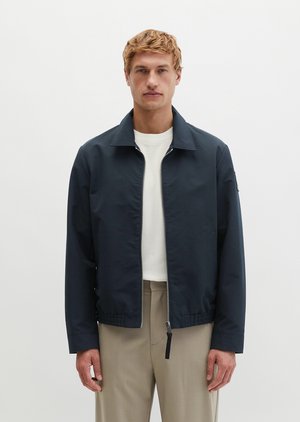 Uomo biondo che indossa una giacca nera con cerniera sopra una camicia bianca e pantaloni beige, in piedi su uno sfondo bianco uniforme.
