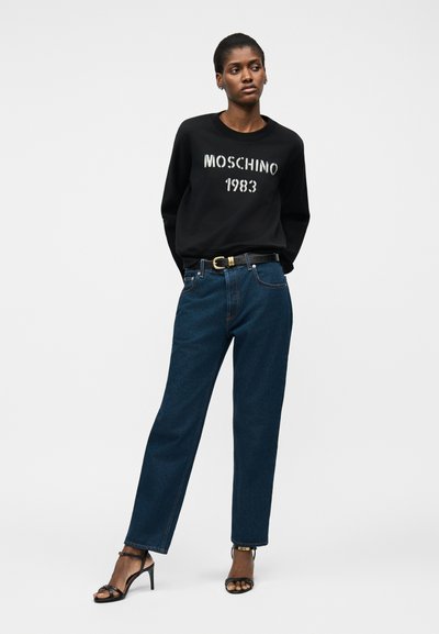 Γυναίκα που φοράει μαύρο φούτερ Moschino 1983, σκούρο μπλε τζιν με μαύρη ζώνη και μαύρα σανδάλια με τακούνι, στέκεται μπροστά σε απλό λευκό φόντο.