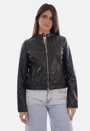 Giovane donna con lunghi capelli scuri che indossa una giacca di pelle nera con zip e jeans azzurri con dettagli di strass.