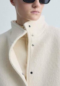 Witte wolmix kraagjas met een gestructureerde afwerking, voorzien van drukknoopsluitingen en een gelaagd ontwerp, gecombineerd met een geribde beige top eronder.