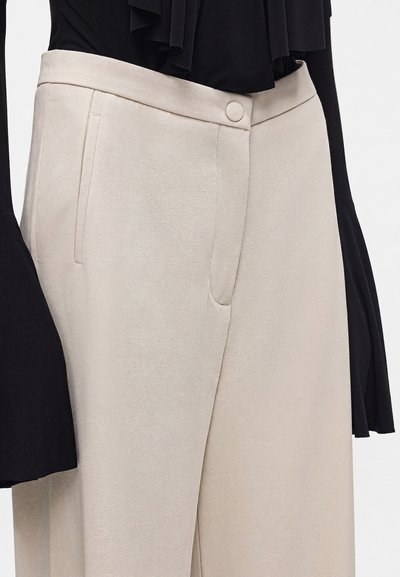 Pantalon beige taille haute avec poches latérales et bouton avant dissimulé, porté avec un haut noir à manches longues avec larges poignets.