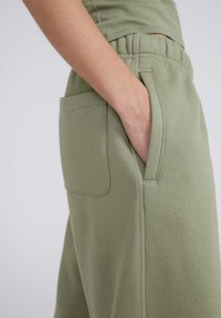 Un pantalon de survêtement vert en coton comporte une poche latérale et une taille élastique. Le tissu a une texture douce et un détail de couture minimal.