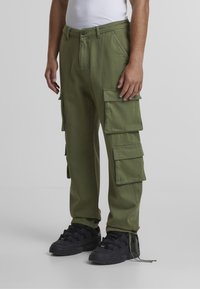 Roheline cargo-püksid, millel on mitu suurt külge taskut, valmistatud vastupidavast kangast; kombineeritud mustade sportlike jalanõude ja valge sporditopiga.