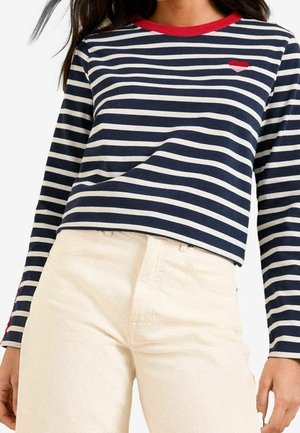 Kobieta w granatowo-białej koszulce z długim rękawem z poziomymi paskami, z czerwonym emblematem serca i czerwonym kołnierzykiem, w połączeniu z beżowymi, wysokim stanem spodniami.