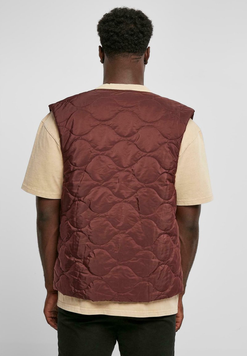 Gilet matelassé bordeaux, présentant un design matelassé avec des motifs circulaires. Le gilet est sans manches et est porté par-dessus une chemise beige clair.