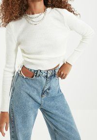 Haut blanc à manches longues côtelé avec des liens sur les côtés et jean denim bleu taille haute. Accents usés et shorts à coupe décontractée.