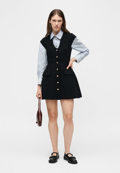 Femme portant une robe noire sans manches boutonnée sur une chemise rayée bleue, tenant un sac à main marron et portant des mocassins noirs.