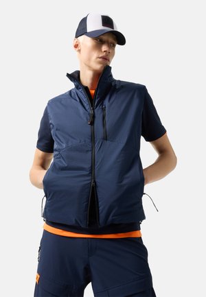 Jeune homme portant une veste zippée bleu marine par-dessus une chemise à manches courtes avec bordure orange, un pantalon cargo bleu marine et une casquette blanche et bleu marine, debout les mains derrière le dos.