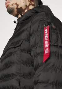 Giubbotto piumino nero con finitura opaca, caratterizzato da trapuntatura orizzontale, cappuccio regolabile con cordino e un'etichetta rossa "Rimuovere prima del volo".