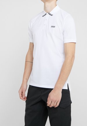 Hombre vistiendo una camiseta pólo blanca de manga corta con cuello de rayas negras y un pequeño logo, combinada con pantalones cargo negros, de pie frente a un fondo gris.