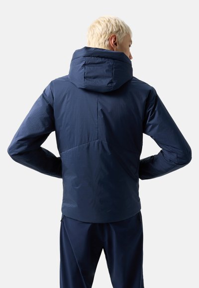 Person with short blond hair wearing a navy hooded jacket and dark pants, facing away against a plain light background.