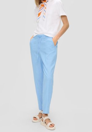 Vrouw draagt lichtblauwe broek met trekkoord, wit kort mouwtjes shirt, gestreepte oranje-blauwe halsdoek en witte gevlochten sandalen.