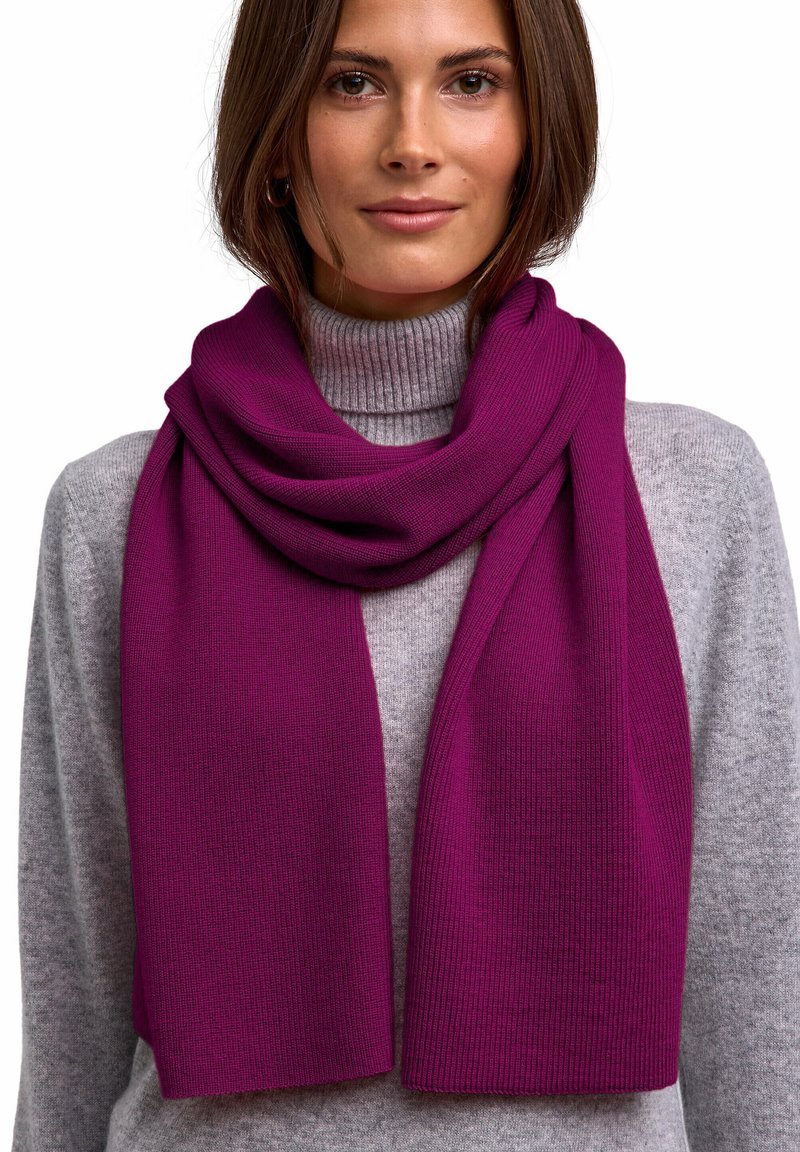 Różowo-fuksjowy dzianinowy szalik o prążkowanej fakturze, luźno ułożony na szarym swetrze z golfem. Charakteryzuje się miękkim, płynnym wzorem z wyraźnymi krawędziami.