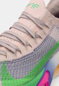 Baskets tricotées rose clair avec des lacets gris, présentant des accents verts et violets, une surface texturée et une semelle en caoutchouc distinctive.