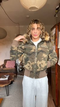 Φόρμα καμουφλάζ με κουκούλα με γούνα, φερμουάρ μπροστά και ριμπ μανσέτες. Φορεμένη με ανοιχτό γκρι φόρμα, σε μια casual εσωτερική σκηνή.