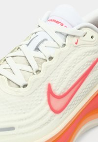 Chaussure de sport blanche avec un swoosh Nike rose vif et des détails orange sur la semelle, avec des lacets blancs et un tissu supérieur texturé.