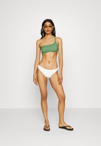 Top de biquíni verde de um ombro só com acabamento trançado, combinado com calções de biquíni brancos. Modelo a usar chinelos pretos e óculos de sol.