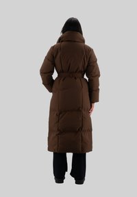 Cappotto imbottito marrone con colletto alto, vita segnato da una cintura e texture trapuntata, che si estende fino a metà polpaccio; indossato sopra pantaloni neri e stivali.