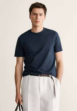 Joven con camiseta azul marino metida en pantalones blancos de cintura alta con un cinturón marrón tejido, sosteniendo una bolsa negra.