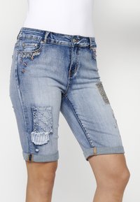 Shorts en denim bleu clair avec des coutures décoratives, divers patchs et des ourlets retroussés. Les détails comprennent des accents brodés et des variations de textile.
