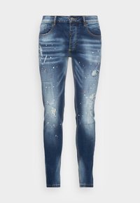 Jeans de mezclilla azul descolorido con parches desgastados, acentos de salpicaduras de pintura, corte recto y diseño clásico de cinco bolsillos.