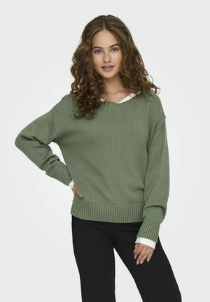 Maglione verde a maglia con scollatura a V, polsini e orlo a coste. Indossato sopra una camicia bianca con colletto. Abbinato a pantaloni neri.