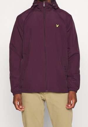 Mann trägt eine bordeauxrote Kapuzenjacke mit Reißverschluss und einem gelben Vogel-Logo, kombiniert mit beigen Hosen, steht vor einem schlichten Hintergrund.