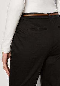 Personne portant un pantalon noir sur mesure avec des poches passepoilées à l'arrière et une fine ceinture marron, associée à un haut blanc à manches longues.