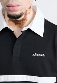 Black polo shirt with a white collar, featuring a front placket and white horizontal stripe. "Adidas" logo in white on the chest area.