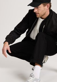 Zwarte bomberjack, witte T-shirt en zwarte broek. Grijze sneakers met witte zolen, witte sokken. Onderwerp zit in een hurkpositie op een lichte achtergrond.