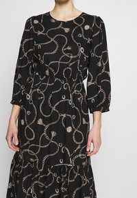 Svart klänning med långa puffärmar, prydd med ett kedje- och tofsmönster i gyllene nyanser. Elastisk midja och design med flerlagad kjol.