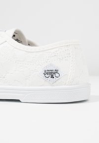 Baskets en dentelle blanche avec un motif floral, semelle en caoutchouc et patch logo décoratif. Texture douce avec des détails cousus le long des bords.