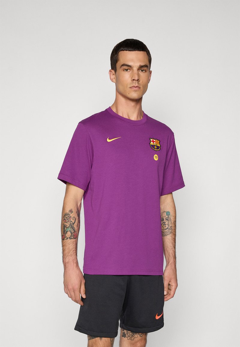 Homem a usar uma t-shirt roxa da FC Barcelona da Nike e calções pretos da Nike, de pé contra um fundo claro liso, com tatuagens nos braços e pernas.