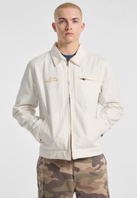 White canvas jacket with a zip closure, stand collar, and left chest pocket. Features "Guess Originals" in gold embroidery.