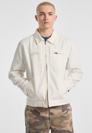 White canvas jacket with a zip closure, stand collar, and left chest pocket. Features "Guess Originals" in gold embroidery.